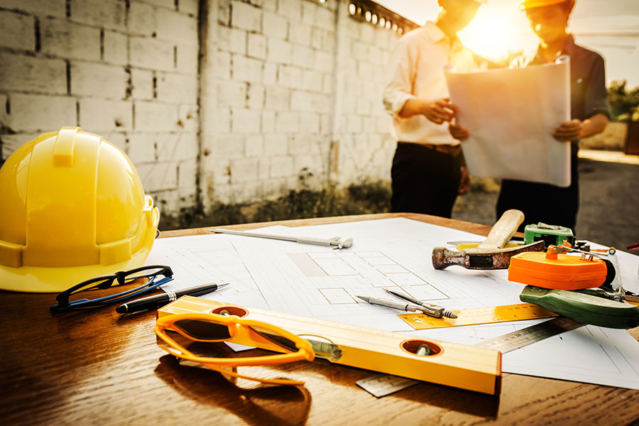 Specialized Business Insurance - Closeup View of Wooden Table With Blueprints and Construction Tools with View of Two Contractors Standing in the Background